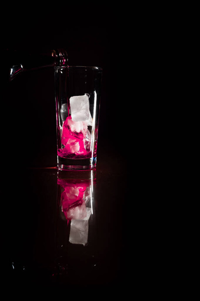 Versare il flusso di soluzione non alcolica rossa sulla parte superiore del ghiaccio nel bicchiere su uno sfondo scuro completo. copiare spazio. Versare alcol, concetto di cantina. - Foto, immagini