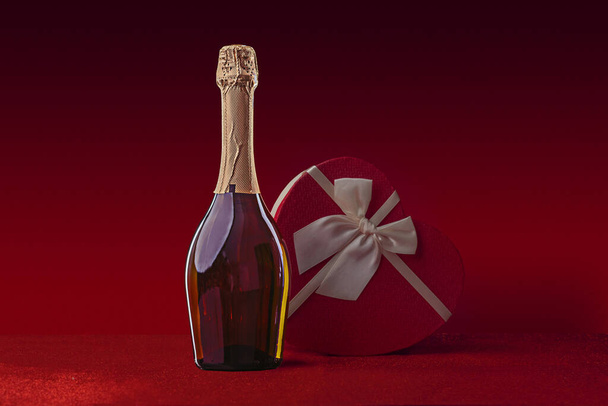 Egy üveg bor és ajándék doboz alakú szív egy íj egy csillogó felületen a piros háttér. Valentin-nap vagy romantikus esti meghívó. - Fotó, kép