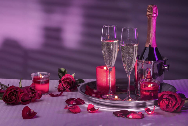 dienblad met kaarsen, een fles in een ijsemmer, glazen mousserende wijn, een decoratie van rode rozen en bloemblaadjes. Wit linnengoed, grijze wand met silhouet of schaduw in paars neon licht. Huwelijksreis, of Valentijnsdag, of een romantische avond, een nacht van lo - Foto, afbeelding