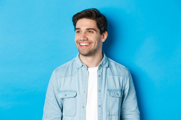 Fiatal modern srác szakállal, balra néz, és mosolyog elégedett fénymásolás tér, felett áll a kék háttér - Fotó, kép