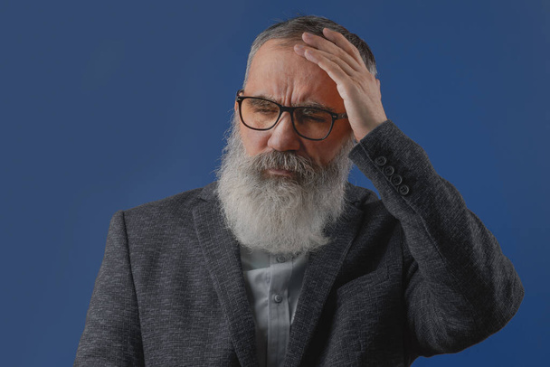 Starszy charyzmatyczny mężczyzna z szarą brodą w jasnej koszuli w ciemnej, luźnej kurtce trzymający głowę, wykazujący migrenę, ból głowy, cierpienie lub zmęczenie. Jasnoniebieskie tło - Zdjęcie, obraz