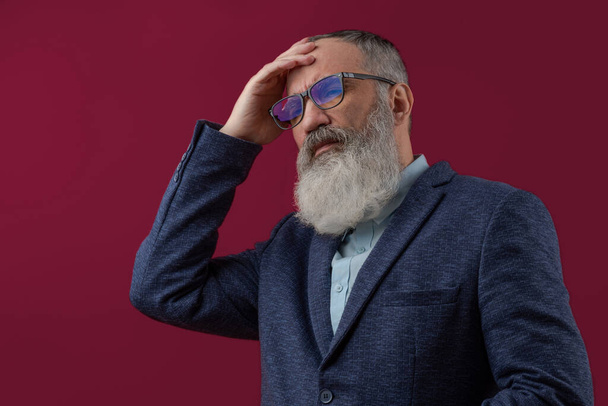 Homme charismatique âgé avec une barbe grise dans une chemise légère dans une main veste décontractée sombre tenant sa tête, montrant migraine, maux de tête, souffrance ou fatigue. Fond rouge vif - Photo, image