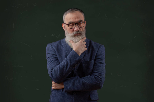 Végzős karizmatikus férfi szürke szakállal, világos pólóban, sötét alkalmi kabát bespectacled kogitative és gondolkodás zöld iskolai tábla háttér - Fotó, kép
