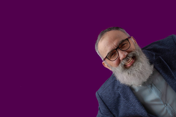 Hombre carismático de risa mayor con una barba gris en una camisa ligera en una chaqueta casual oscura lleva gafas. Fondo magenta brillante - Foto, Imagen