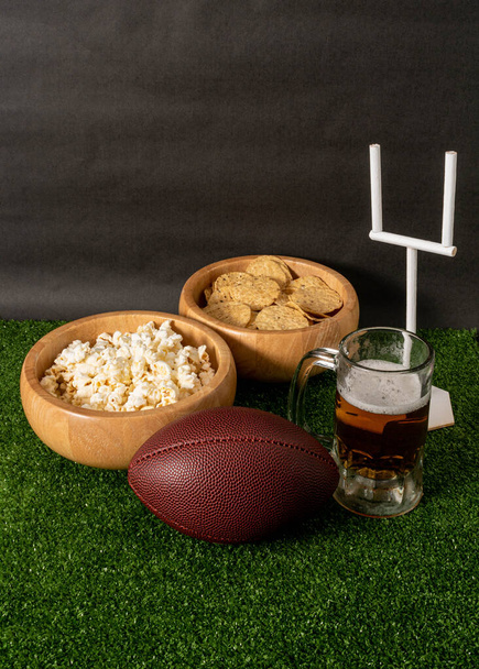 Американская футбольная вечеринка. Американский футбольный мяч, пиво, закуски и гол пост на зеленой траве и черном фоне - Фото, изображение