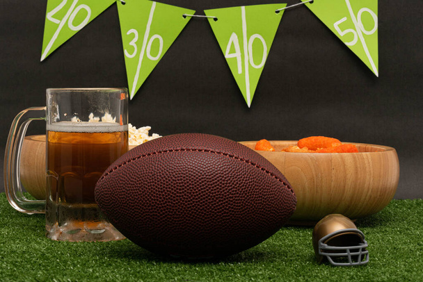 Fiesta de fútbol americano. Pelota de fútbol americano, cerveza, casco de oro, bocadillos y pancarta sobre hierba verde y fondo negro - Foto, imagen