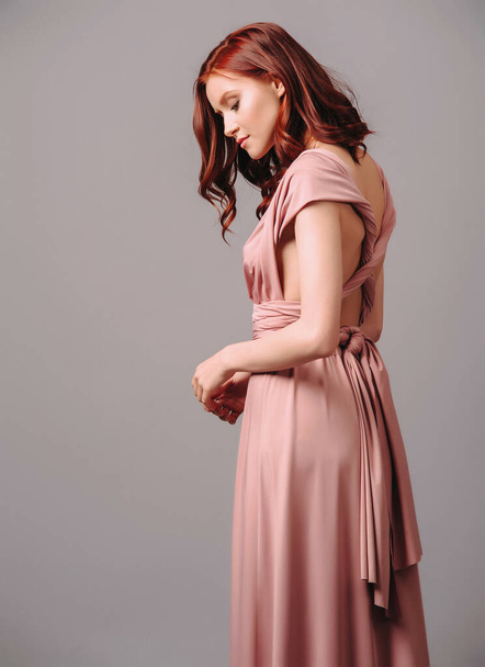 Bridesmaid's look in dust pink. Young ginger woman in summer transformer evening dress on grey background at studio. Gorgeous sexy lady. - Photo, Image