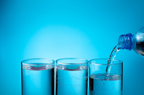 Eau dans des verres, remplissage de trois verres à partir d'une bouteille en plastique sur un fond bleu. - Photo, image