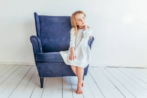 Süßes kleines Mädchen in weißem Kleid sitzt auf einem modernen gemütlichen blauen Stuhl und entspannt es sich im weißen, hellen Wohnzimmer zu Hause drinnen. Kindheit Schüler Jugend entspannen Konzept. - Foto, Bild