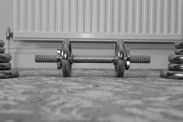 Fekete-fehér fénykép egy kis súlyzóról otthon a padlón, nagyobb súllyal mindkét oldalon. - Fotó, kép