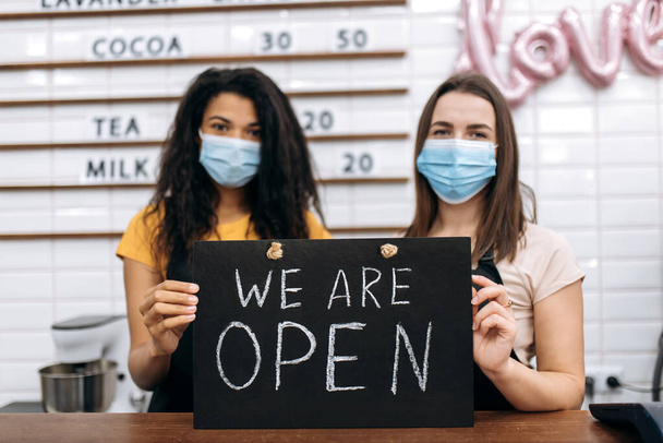 Дві роздягнені офіціантки, кав "ярня або ресторан, афроамериканська та кавказька, стоять з підписом" МИ Є ОПЕН "за прилавком бару з медичними масками - Фото, зображення