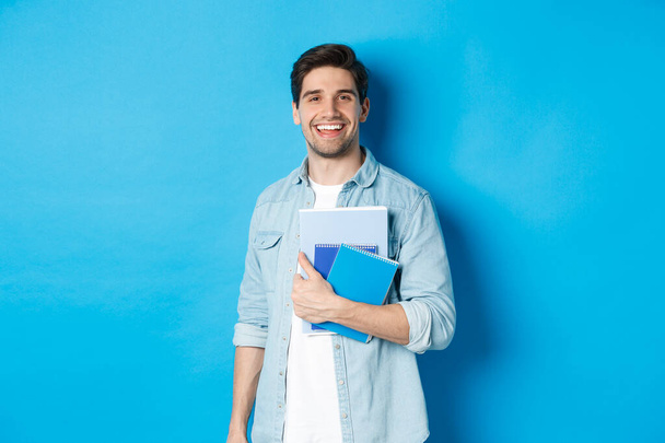 Smiling man studying, holding notebooks and looking happy, standing over blue background - Photo, Image