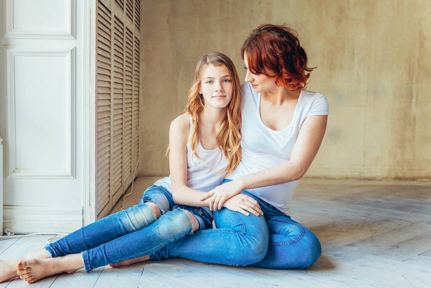 Young mother embracing her child. Woman and teenage girl relaxing in white bedroom near gray wall indoors. Happy family at home. Young mom playing with her daughter. - Photo, Image