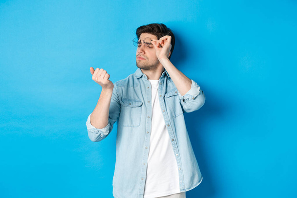 Jonge knappe man met een bril die naar zijn nagels kijkt, manicure controleert, over een blauwe achtergrond staat - Foto, afbeelding