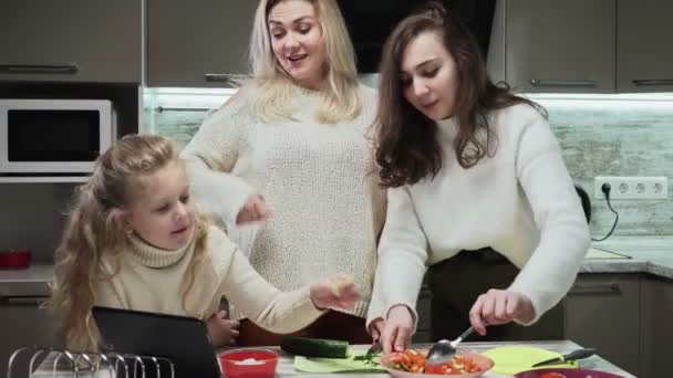 Jonge gelukkige familie, moeder en twee haar dochters koken salade in de keuken. Moeder gebruikt tablet met recept van smakelijke salade met tomaten en komkommers - Video