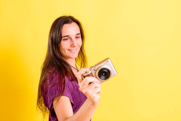 Young photography student girl holds a compact camera, isolated on yellow studio background. - Photo, Image