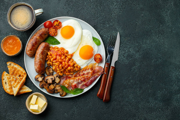 Повний англійський сніданок на тарілці з смаженими яйцями, ковбасою, беконом, бобами, тостами і кавою на темному кам'яному тлі. Простір копіювання. Краєвид - Фото, зображення