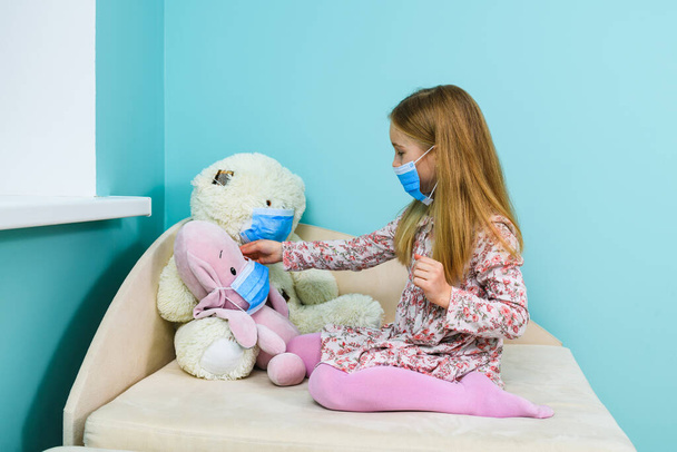 Une petite fille joue avec un ours en peluche et des jouets en peluche de porc pendant la quarantaine à domicile et les études à distance, portant trois masques chirurgicaux bleus - Photo, image