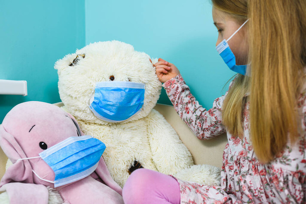 Girl play and care about stuffed toys during quarantine, wearing protective blue disposable masks at blue indoor background - Photo, Image