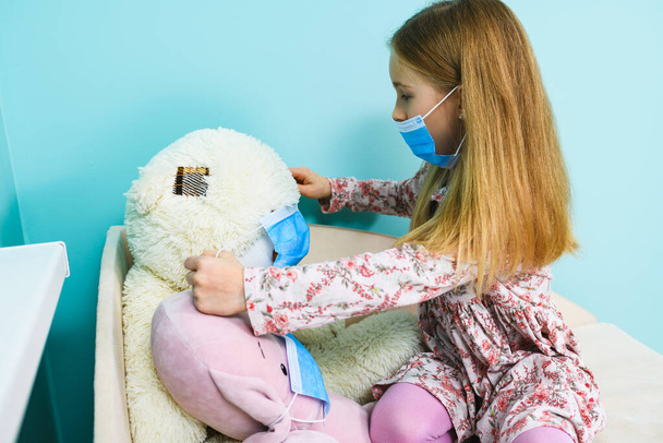 Small girl play at home during lockdown isolation, wearing blue surgical face mask on herself and stuffed toys - Fotografie, Obrázek