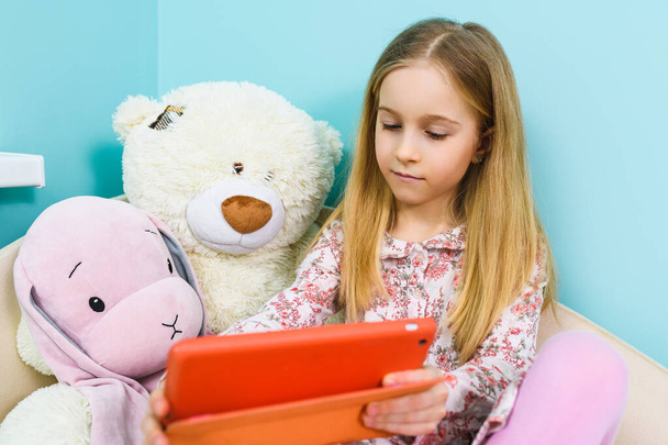 Small girl primary school age sit on sofa with teddy bear and looking at tablet screen inside light pastel home interior during lockdown isolation - Photo, Image