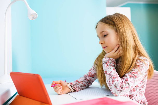 Kind im Grundschulalter sitzt mit leerem Schulheft am Schreibtisch, hält Stift in der Hand und schaut auf Tablet-Bildschirm, lernt während der Sperrung zu Hause - Foto, Bild