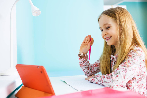 Μικρό κορίτσι του σχολείου χαμογελώντας και χαιρετισμό στην οθόνη tablet κατά τη διάρκεια απομακρυσμένης περιόδου απομόνωσης μελέτη καραντίνα, κοινωνική απόσταση - Φωτογραφία, εικόνα