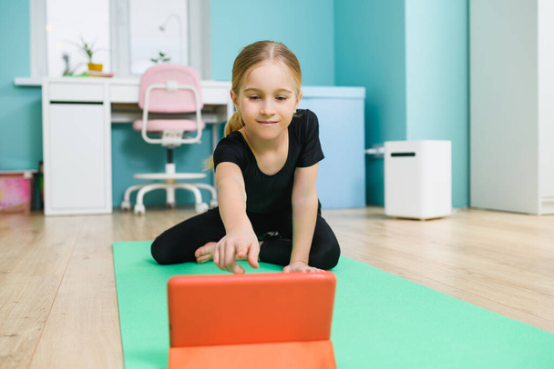 Exercício positivo menina da escola em casa online com tablet, situando-se no tapete esporte em casa interior durante a quarentena - Foto, Imagem
