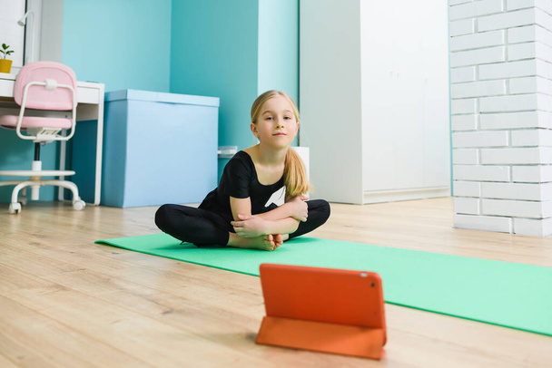 小学生の女の子はスポーツマットとタブレットを床に置きます,黒のレオタードを身に着けています前に開始オンラインリモート体操教室中に自宅で隔離 - 写真・画像