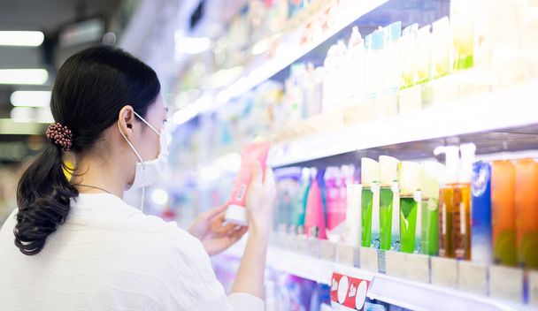 Cerrahi maske takan Asyalı kadın ve süpermarketteki cilt bakımı koridorunda alışveriş yapmak için yüz köpüğü şişesini tutan Covid-19 salgını, alışveriş konsepti, . - Fotoğraf, Görsel