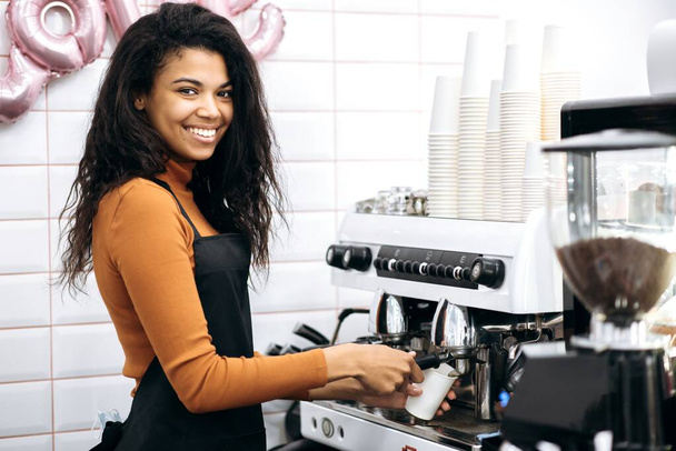 Oldalnézet egy mosolygós barátságos afro-amerikai nő barista csinál kávét papírpohárban a kávézó látogató a kávéfőző gép, áll a pult mögött, és mosolyog - Fotó, kép