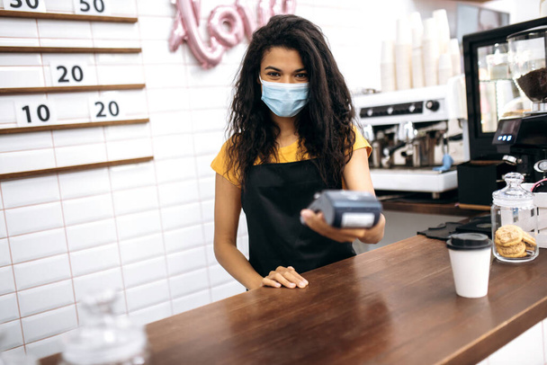 Afroamerikanische Barista, Kellnerin oder Kleinunternehmerin mit medizinischer Schutzmaske, die dem Besucher ein kontaktloses Zahlungsterminal bietet - Foto, Bild