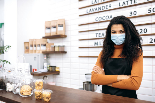 Portrait of a pretty waitress wearing uniform and protective medical mask. Young African American female barista stands behind the bar counter and looks at the camera. Small business concept - Foto, Imagen