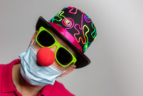 Man with medical mask, clown nose, sunglasses and colorful hat at carnival in covid-19 and corona times with copy space. Horizontal - Photo, Image