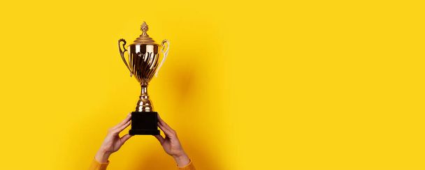 Raised hands of a woman are holding trophy over yellow background - Photo, Image