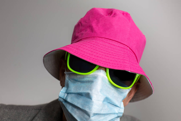 Porträt eines anonymen Mannes mit Chirurgenmaske, gelber Sonnenbrille und rosa Hut im Profil zu Zeiten von Covid 19 und Corona. Hochwertiges Foto - Foto, Bild