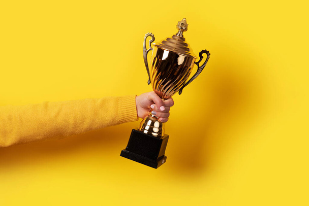 рука тримає золотий трофей над жовтим тлом
 - Фото, зображення