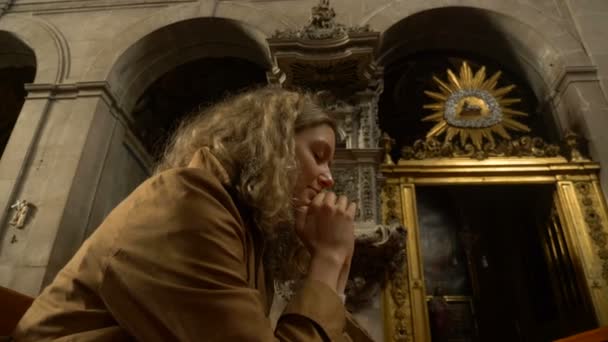fiel mujer reza en la iglesia católica con los ojos cerrados sentado en el banco - Imágenes, Vídeo