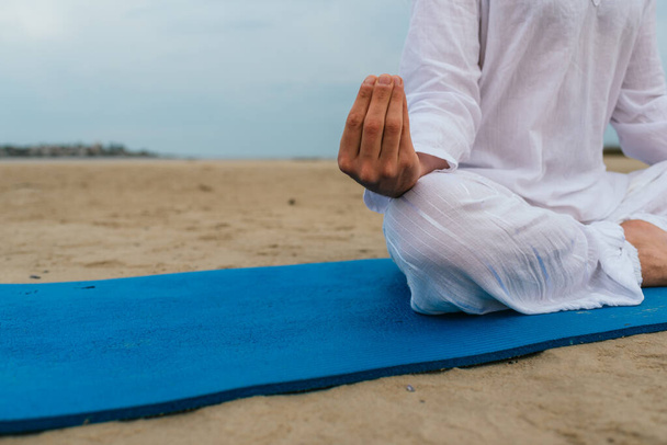 Close up de homem não reconhecido sentado no tapete de ioga na praia fazendo poses de ioga. - Foto, Imagem