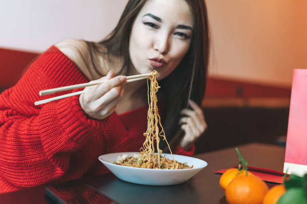 Prachtige glimlachende jonge Aziatische vrouw in rode kleren noedels etend met bamboe eetstokjes in het Chinese Vietnamese restaurant - Foto, afbeelding