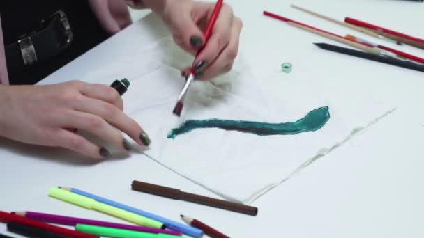 Atraktivní mladá žena kreslí zelenými akrylovými barvami na bílou látku u stolu - Záběry, video