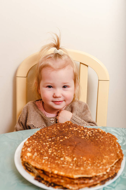 A beautiful little girl sits at a table with large round pancakes. Carnival festival. - 写真・画像