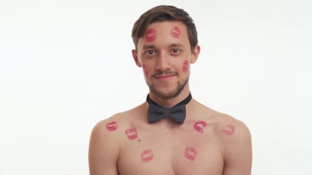 Close up portrait of Caucasian beautiful young brunette bearded man shirtless wear grey bow tie with red lipstick on body and head blowing kiss isolated on white wall background. Women day concept. - Footage, Video