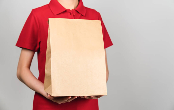 Empleada mujer en camisa roja uniforme sosteniendo paquete de papel artesanal aislado sobre fondo gris. - Foto, imagen