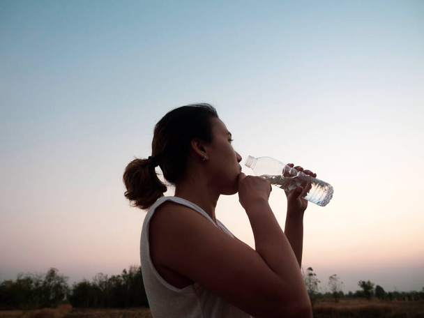Azjatka pijąca wodę z plastikowej butelki po treningu wieczorem latem na pięknym tle zachodu słońca. Koncepcja sprawności i zdrowego stylu życia. - Zdjęcie, obraz