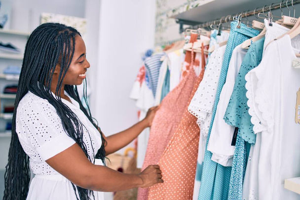 Joven afroamericana americana sonriendo feliz sosteniendo ropa en tienda de ropa - Foto, imagen