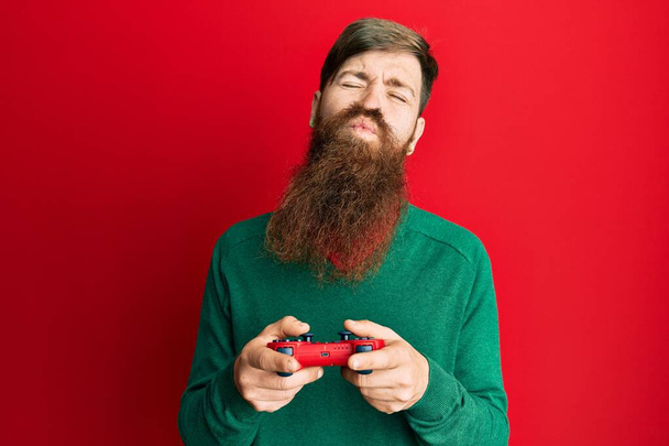 Redhead man with long beard playing video game holding controller looking at the camera blowing a kiss being lovely and sexy. love expression.  - Foto, Bild