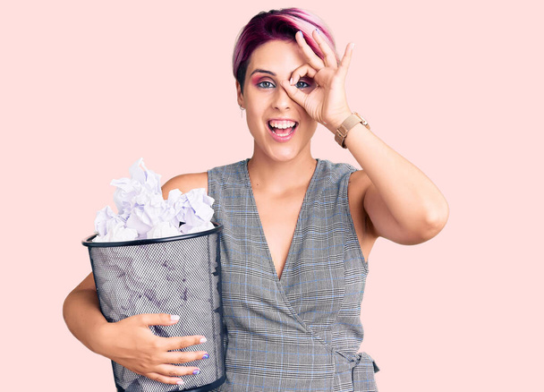 Jonge mooie vrouw met roze haar vasthouden papieren bak vol met verfrommelde papieren glimlachen gelukkig doen ok teken met de hand op oog kijken door vingers  - Foto, afbeelding