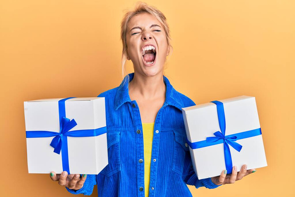 Young blonde girl holding gifts angry and mad screaming frustrated and furious, shouting with anger looking up.  - Photo, Image
