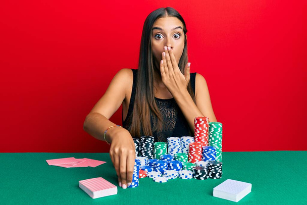 Young hispanic woman sitting on the table holding casino chips covering mouth with hand, shocked and afraid for mistake. surprised expression  - Photo, Image